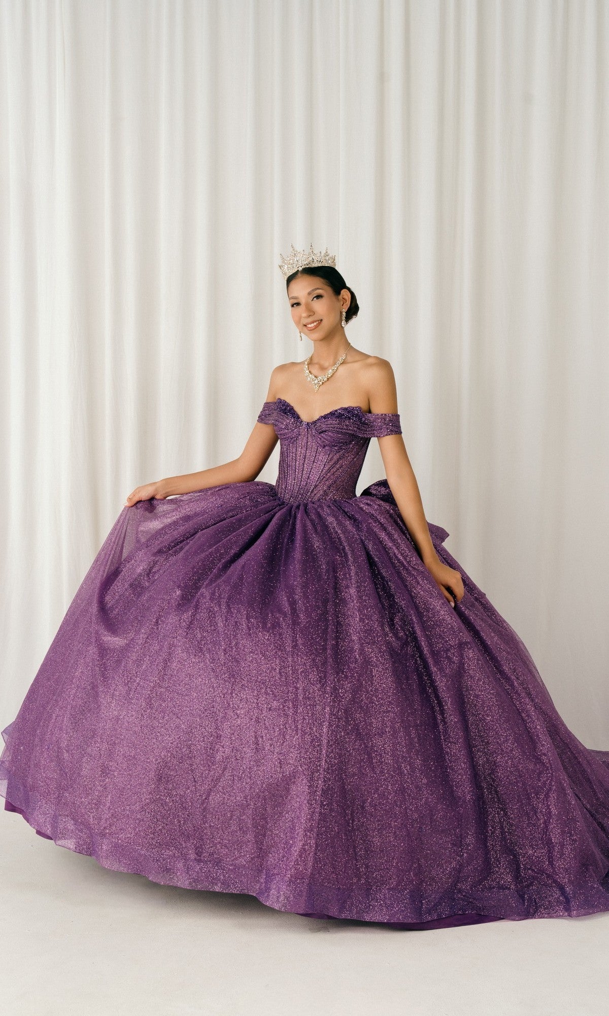 A woman stands before white draped curtains, wearing a sparkling purple Quinceanera Dress 8460JQ By Cinderella Couture and a tiara.